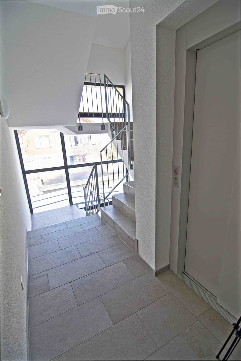 Modern hallway with tiled flooring, white walls, and a staircase leading upwards with a metal railing and glass balustrade. Natural light streams in from a large window at the top of the stairs.