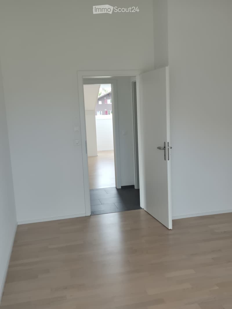 Empty hallway with light-colored wooden flooring and white walls, featuring an open doorway leading to another room.