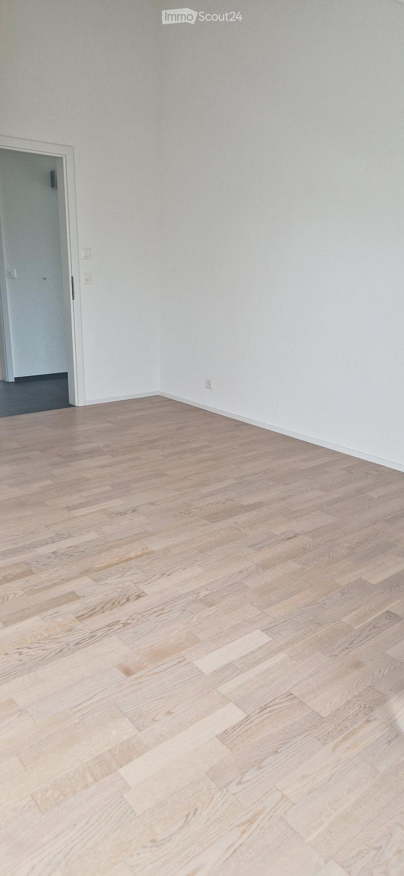 Empty room with light-colored wooden flooring and white walls. A doorway leads to another room, and electrical outlets are visible on the walls.
