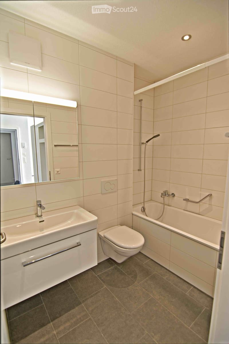 Modern bathroom with white tiles, a sink, toilet, bathtub, and a large mirror with built-in lighting.