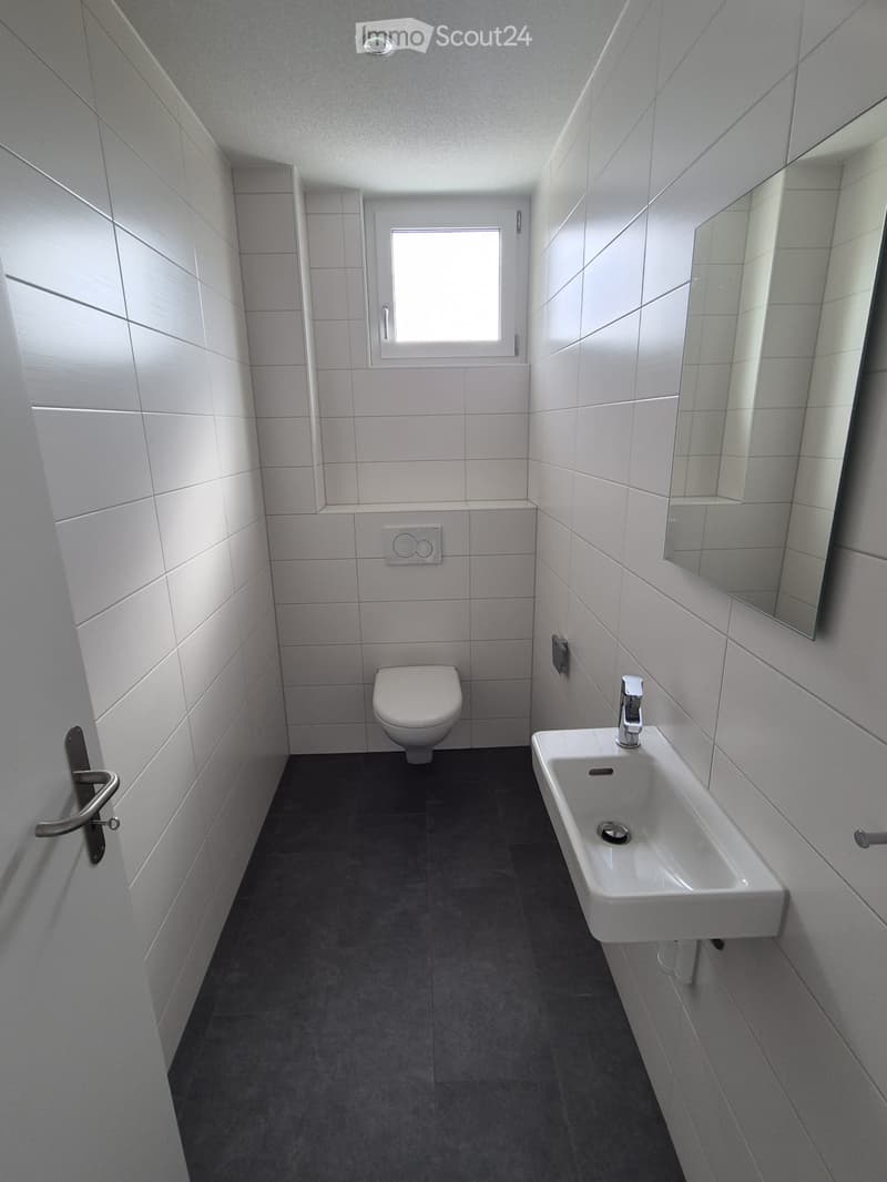 Modern bathroom with white tiled walls, a toilet, a sink with a chrome faucet, and a large mirror. The floor is dark grey tiles, and there is a small window above the toilet.