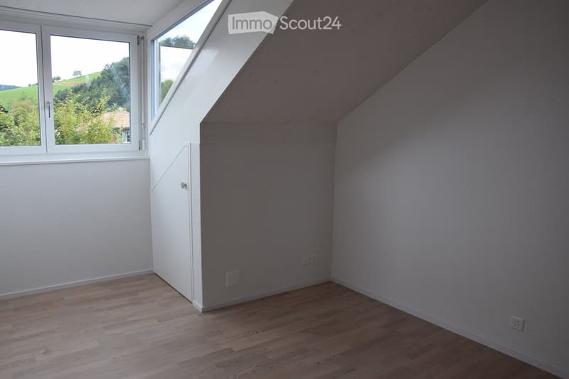 Empty attic room with wooden flooring, white walls, and large windows offering a view of greenery outside.