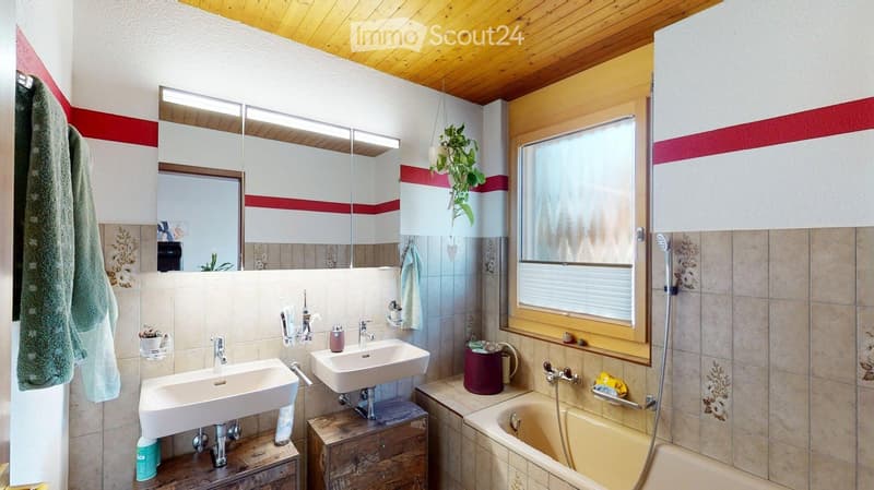 Bathroom with two sinks, a bathtub, a large mirror, and a window with blinds. The walls are tiled, and there are towels and a plant visible.