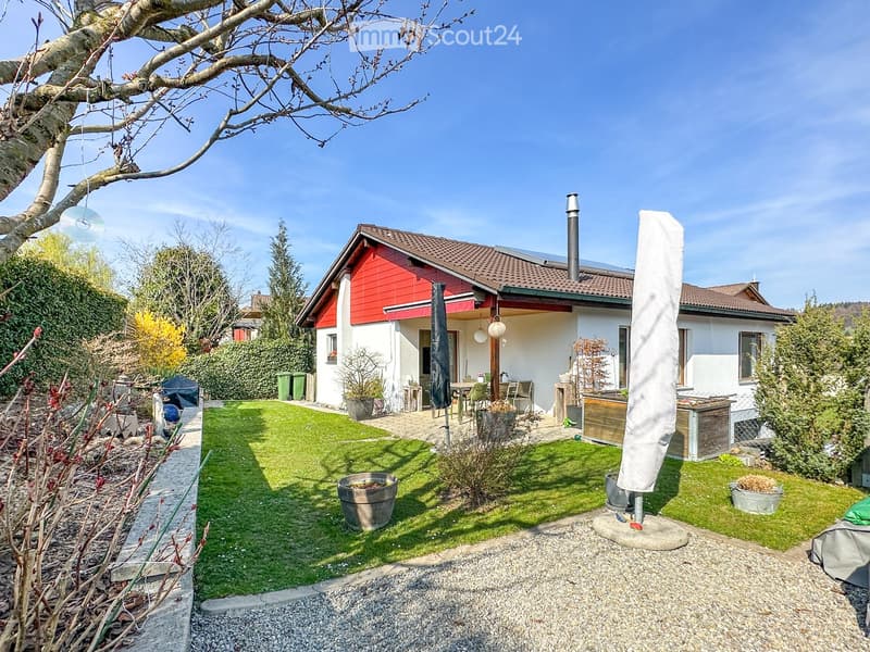 Well-maintained garden with a lawn, patio seating area, outdoor umbrella, and surrounding greenery.