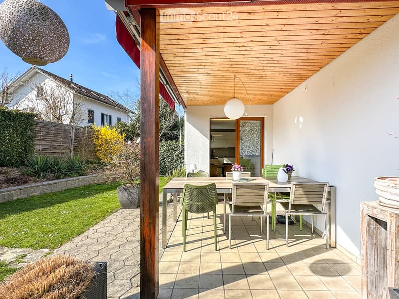 Outdoor patio with a wooden pergola, tiled floor, dining table with chairs, and a view of a well-maintained garden.