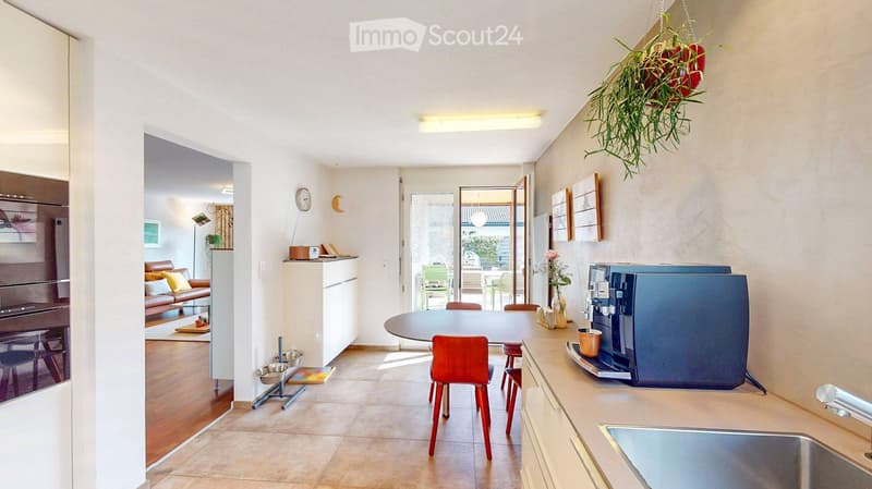 Open-plan kitchen and living area with modern furniture, including a round dining table with chairs, a coffee machine, a sofa, and a refrigerator. The space features light-colored walls, wooden flooring, and a hanging plant.