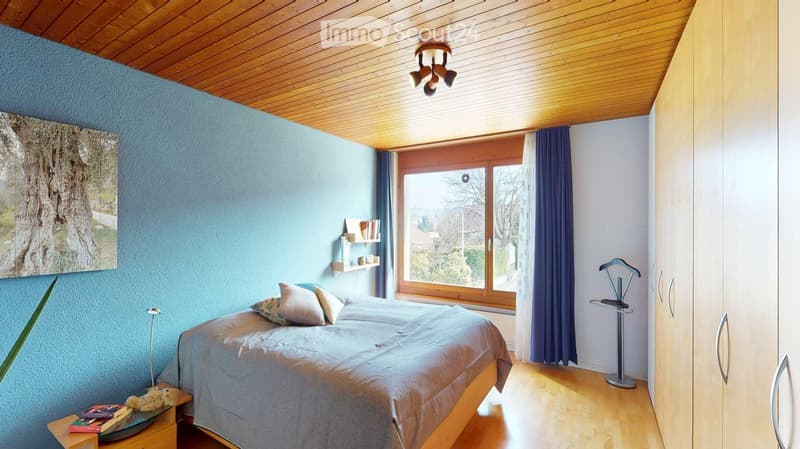 Cozy bedroom with wooden ceiling, blue walls, large window with blue curtains, bed with pillows and duvet, nightstand with books, picture frame, and a coat rack.