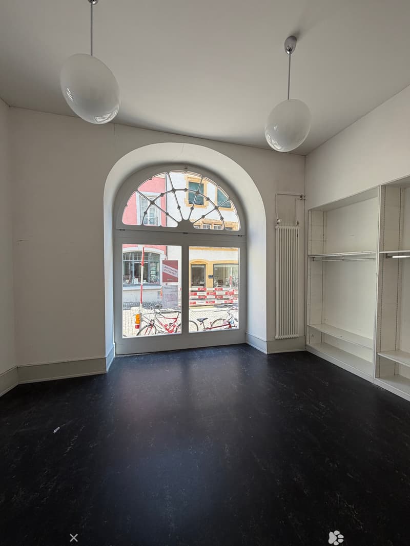 Empty room with dark flooring, white walls, large arched window, and built-in shelving unit. No furniture or equipment present.