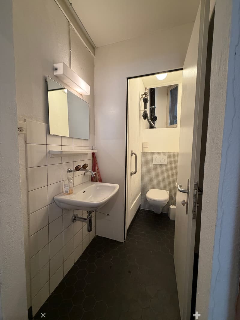 Modern bathroom with a white sink, mirror, and toilet. Features hexagonal dark tiles on the floor and white tiled walls.