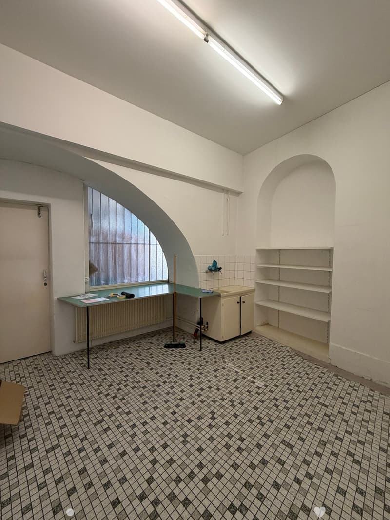 Small room with tiled floor, white walls, a table with papers, a sink, a cabinet, and shelves.