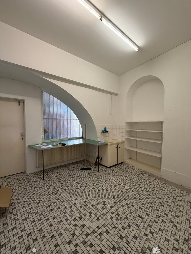 Small room with tiled floor, white walls, a table with papers, a sink, a cabinet, and shelves.
