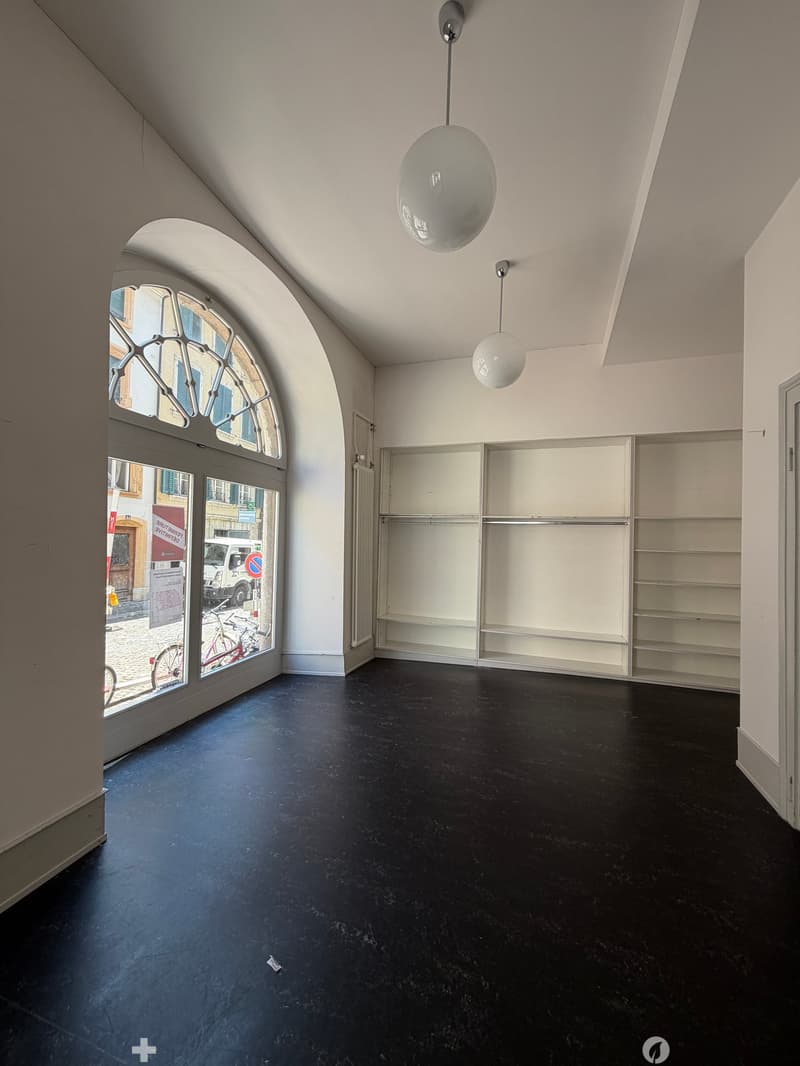 Empty room with dark flooring, white walls, built-in shelving unit, and large arched window with glass panes. Two modern pendant lights hang from the ceiling.