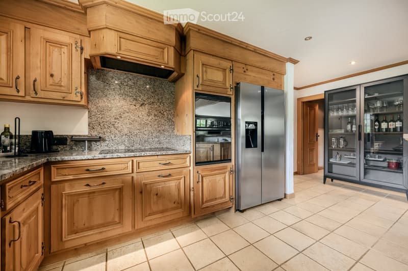 A well-furnished kitchen with wooden cabinets, granite countertops, a stainless steel refrigerator, and a wine rack. The space features a modern design with ample storage and a clean, organized layout.