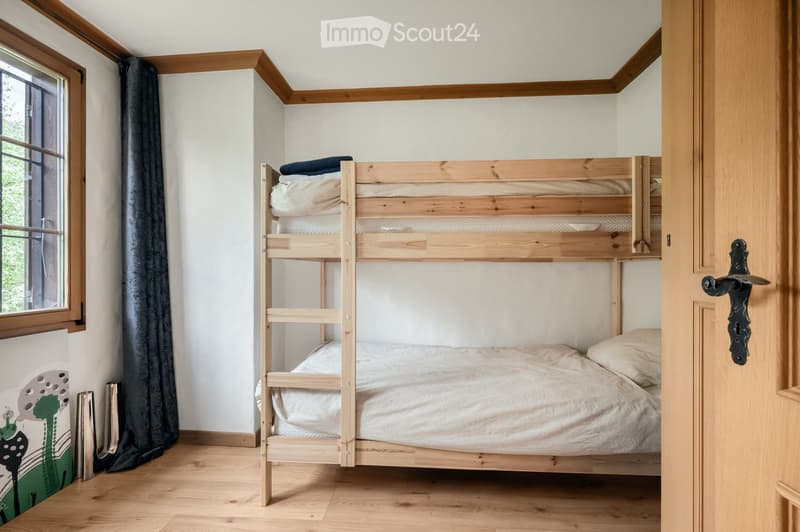 A small bedroom with a wooden bunk bed, neatly made with pillows and blankets, and a window with dark curtains.
