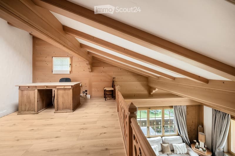 Spacious, wooden-furnished room with exposed wooden beams, a large window, a sofa, a small table, a desk, and a chair.
