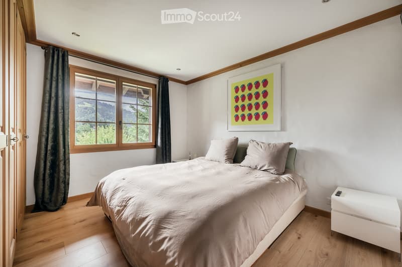 Cozy bedroom with wooden floors, large window with mountain view, bed with beige bedding, nightstand, and a colorful fruit-themed painting on the wall.