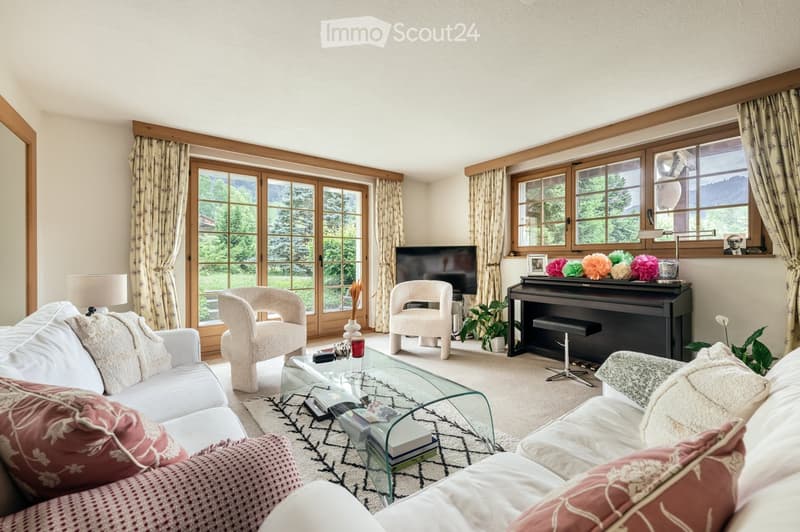 A well-lit living room with large windows and wooden frames, featuring a white sofa with red cushions, a glass coffee table, armchairs, a piano, and decorative items on the windowsill.