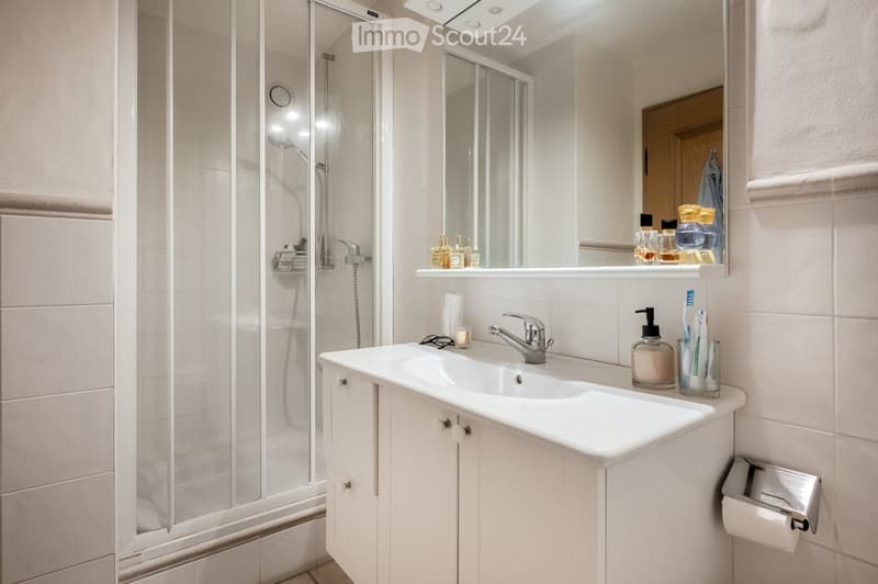 Modern bathroom with white tiles, a large sink with a modern faucet, a mirror with cosmetics on the shelf, a glass-enclosed shower, and a toothbrush holder with toothbrushes and a soap dispenser.