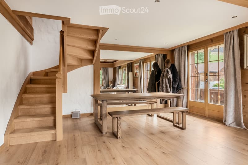 Spacious, light-filled room with wooden flooring, a large wooden table and benches, a staircase leading upwards, large windows with curtains, and a mirror on the wall.