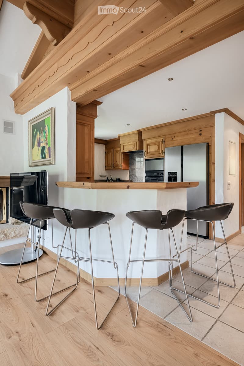 Modern kitchen with wooden bar counter, four black stools, stainless steel appliances, and exposed wooden beams on the ceiling.