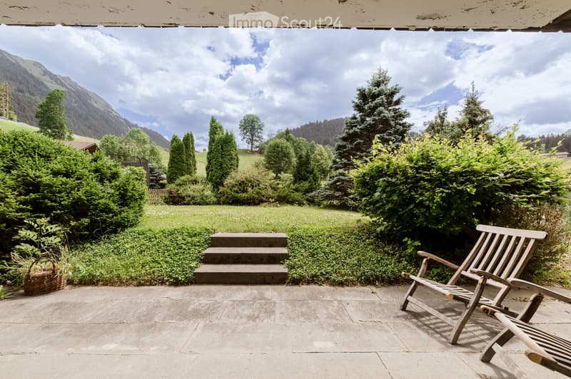 A serene garden area with a stone-paved patio, wooden lounge chairs, a small basket, and lush greenery, overlooking a picturesque mountain landscape.