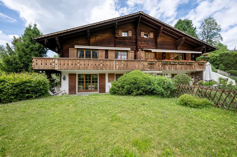A two-story wooden house with a large balcony, surrounded by a well-maintained lawn and garden, with trees and shrubs in the background.