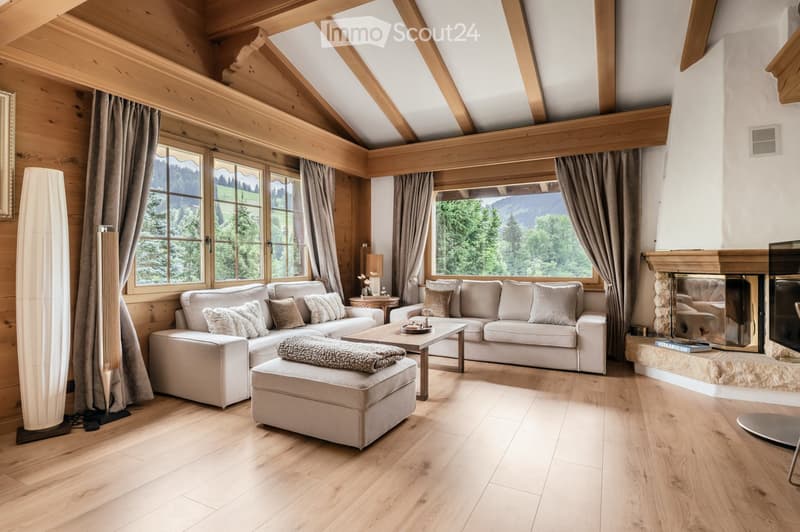 Cozy living room with exposed wooden beams, large windows offering a scenic mountain view, a fireplace, a sectional sofa, an ottoman, a coffee table, and a television.