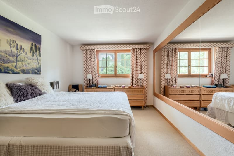 Cozy bedroom with a large bed, nightstands, lamps, a large mirror, and a scenic painting on the wall. The room features wooden furniture and patterned curtains.