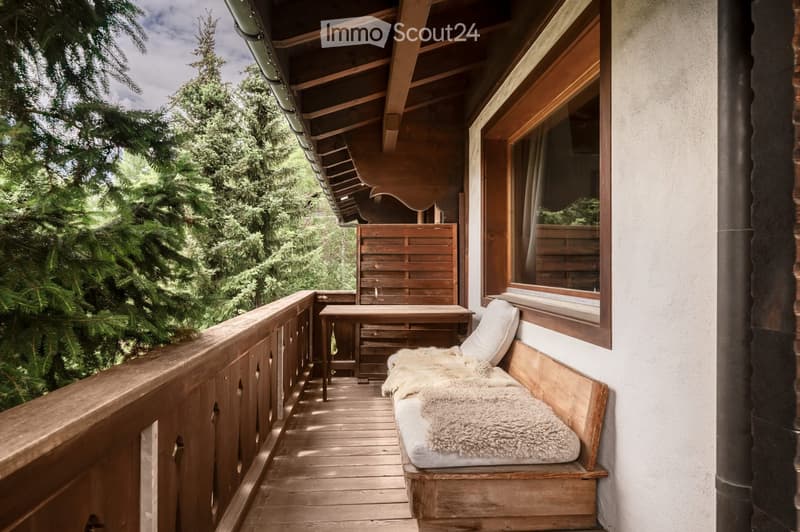 Wooden balcony with a bench, small table, and chair, surrounded by lush greenery.