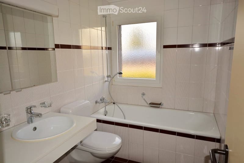 A modern bathroom with white tiles, a bathtub, toilet, sink, and a large mirror. The room features a frosted window and a handheld showerhead.