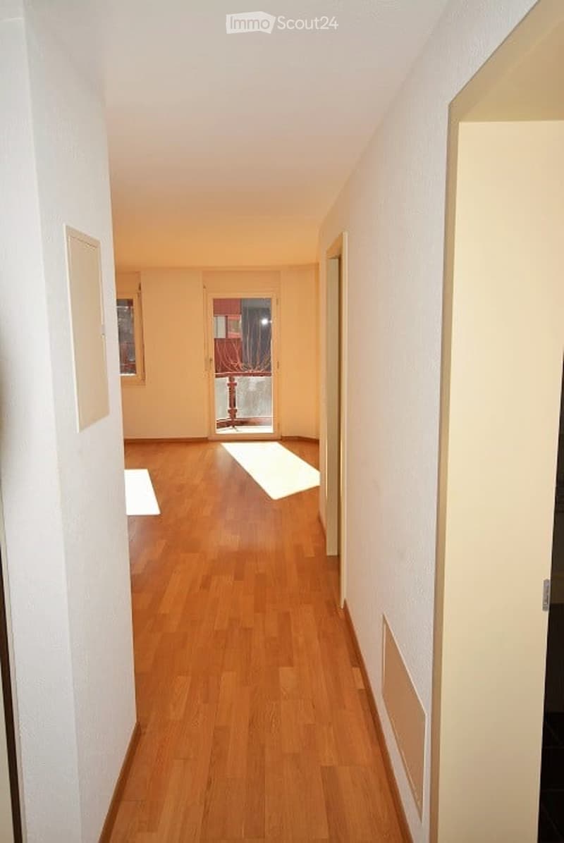 A long, narrow hallway with light wooden flooring and white walls. The hallway features several doors and a small window at the far end, allowing natural light to enter.