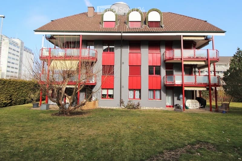 Multi-story apartment building with red shutters, balconies, and a well-maintained lawn in front.