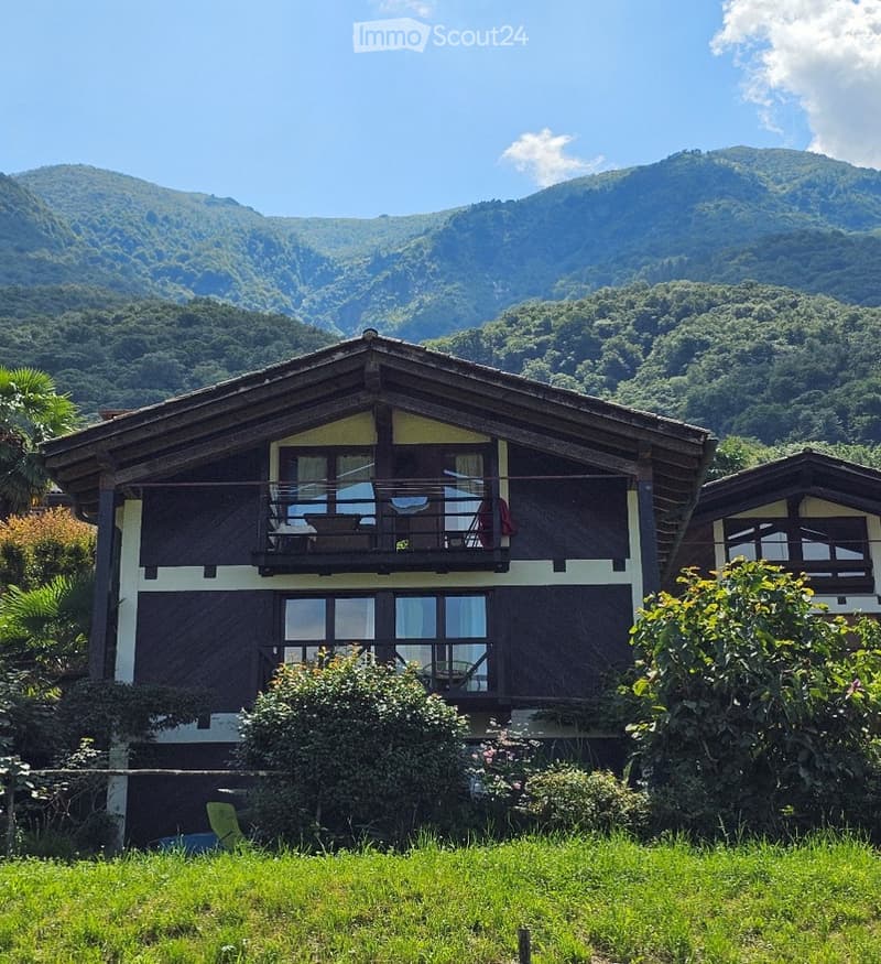 Two-story wooden house with large windows and a balcony, surrounded by lush greenery and mountains.