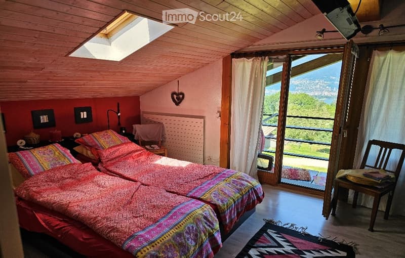 Cozy attic bedroom with slanted wooden ceiling, skylight, two single beds with colorful bedding, nightstand, chair, and a view of the landscape through large windows.