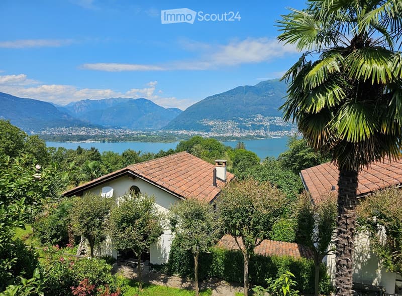 A serene garden view with a small white house, surrounded by lush greenery and palm trees, overlooking a lake and distant mountains.