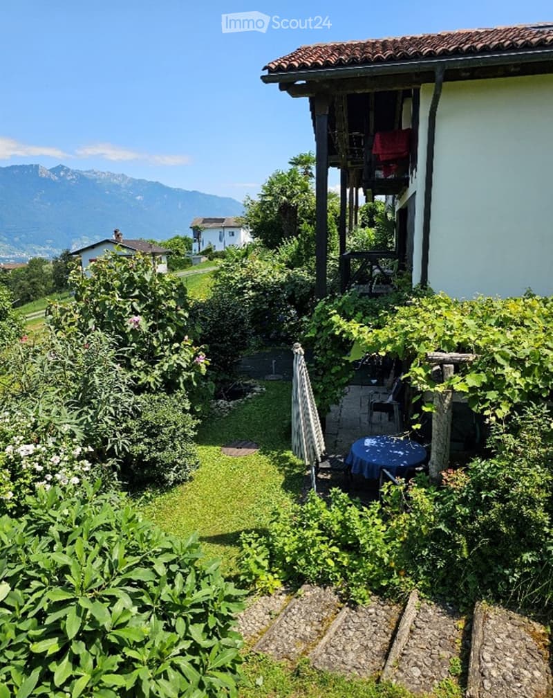 Garden area with a small patio, table, and chairs, surrounded by greenery and offering a scenic mountain view.