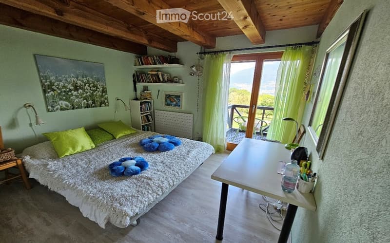 Cozy bedroom with a large bed, green pillows, a white fur throw, a desk with office supplies, a bookshelf, and a view of the landscape through large windows.