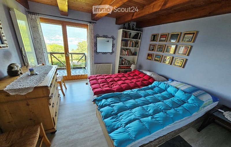 Cozy bedroom with wooden beams, large window with balcony access, double bed with blue and red duvet, nightstand, bookcase, mirror, and small table.