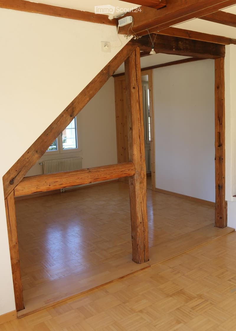 Empty room with wooden floors, exposed wooden beams, and a staircase leading to another level. There are windows and a radiator visible.