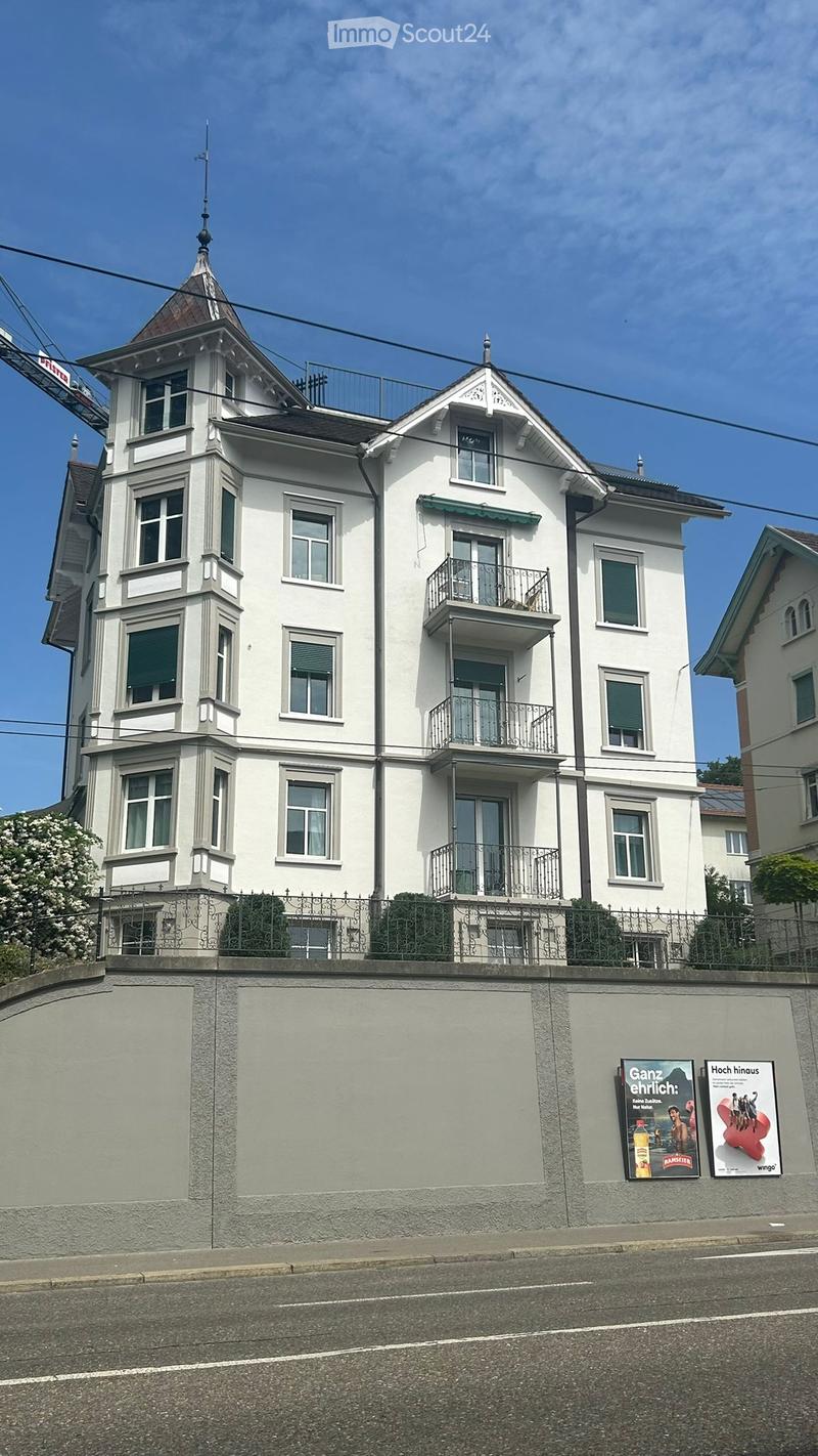 A multi-story white residential building with balconies, decorative roofline, and ornate railings. The building has multiple windows and is surrounded by a low wall with posters on it.
