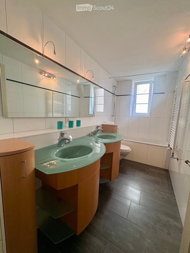 Modern bathroom with double sink, large mirror, bathtub, and toilet. Features wooden cabinets and green countertops.