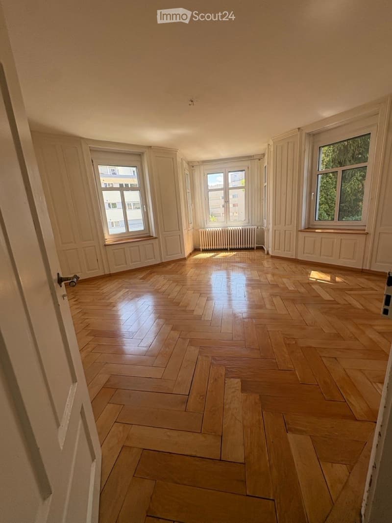 Empty room with wooden parquet flooring, white walls, and large windows with wooden sills. Natural light illuminates the space, and there is a radiator under one window.