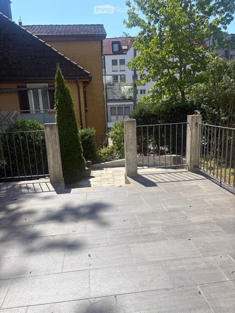 A paved outdoor area with a metal fence, stone pillars, and surrounding greenery, including trees and shrubs.