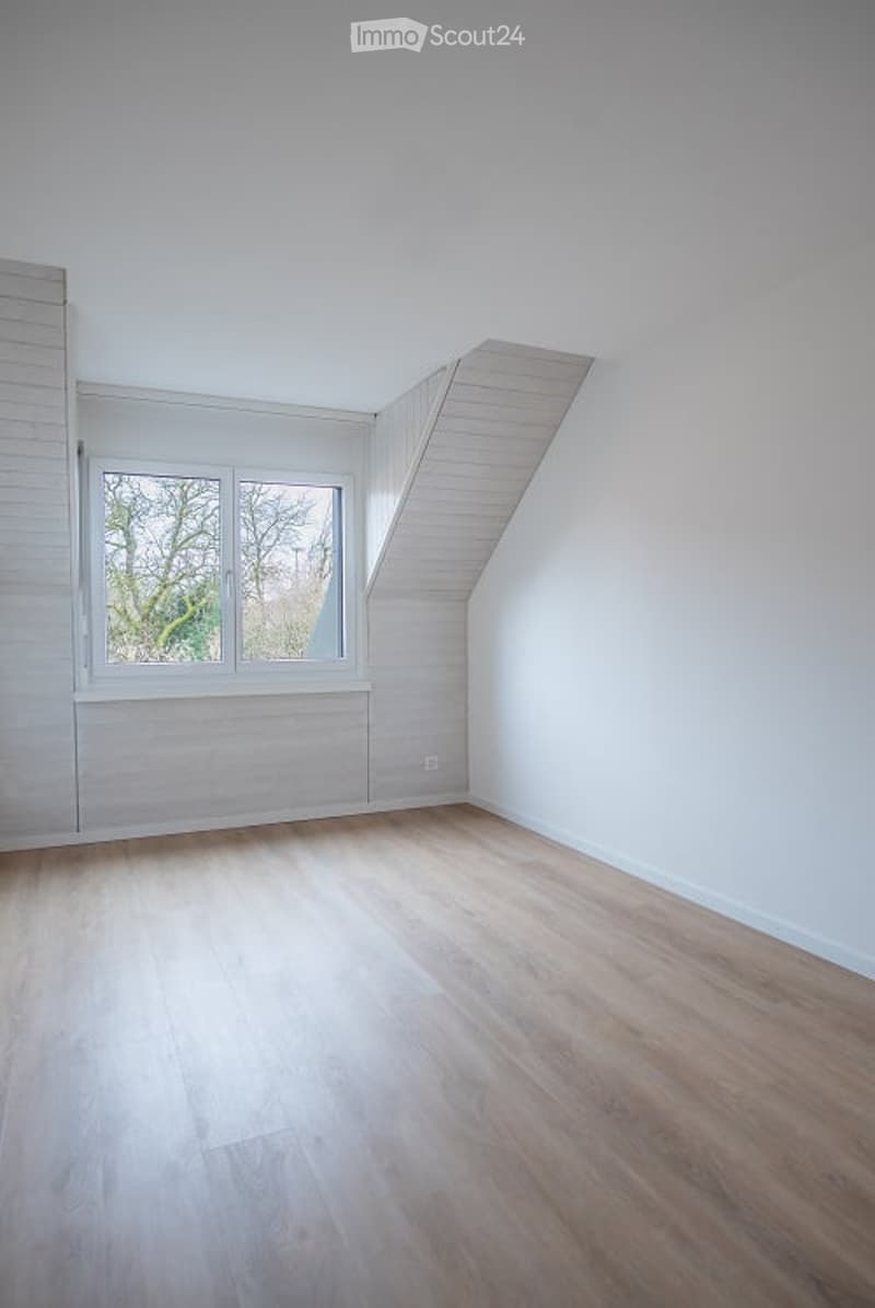 Completely empty room with white walls, light wooden flooring, and a large window with white frames. The room has a slanted ceiling typical of an attic space.