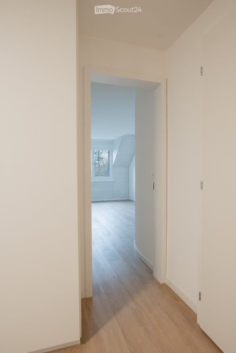 Empty room with light wooden flooring and white walls, featuring an open doorway leading to another room with a skylight window.