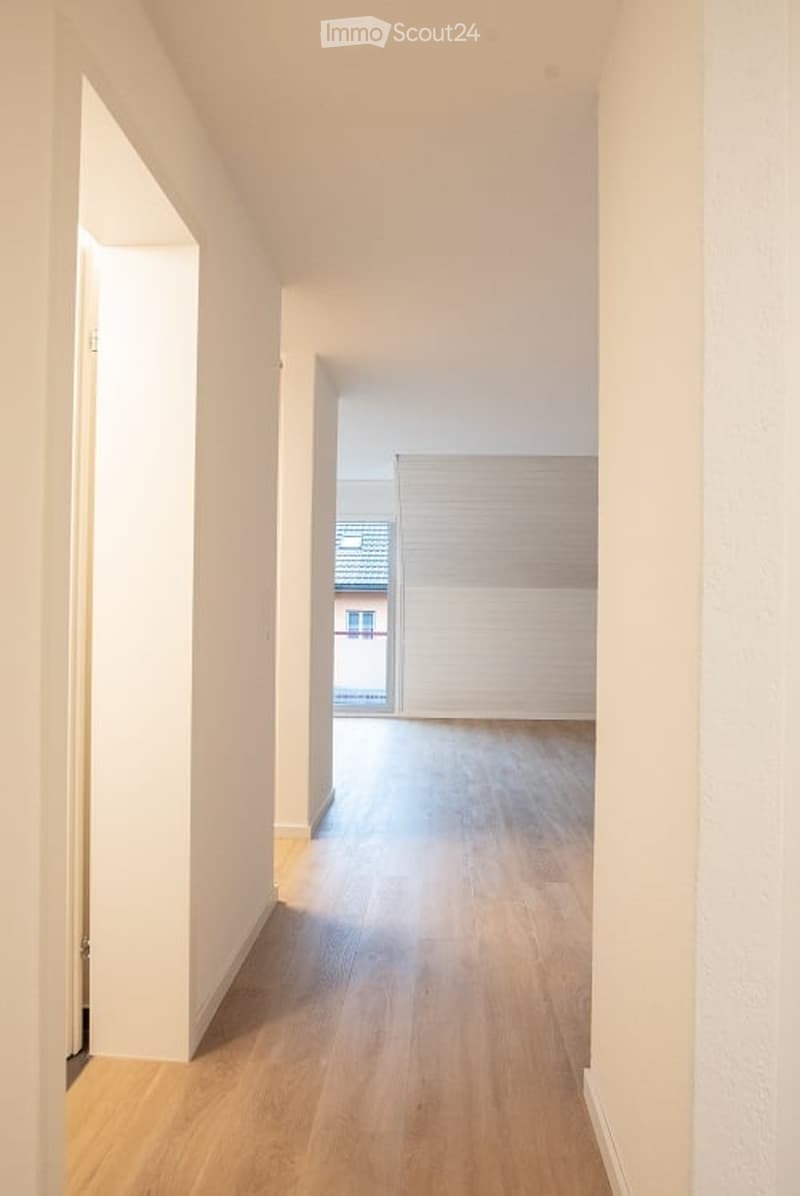 Empty hallway with light wooden flooring and white walls, leading to an open doorway.