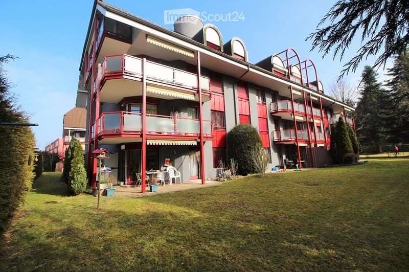 Multi-story apartment building with red and white balconies, surrounded by green lawn and trees.