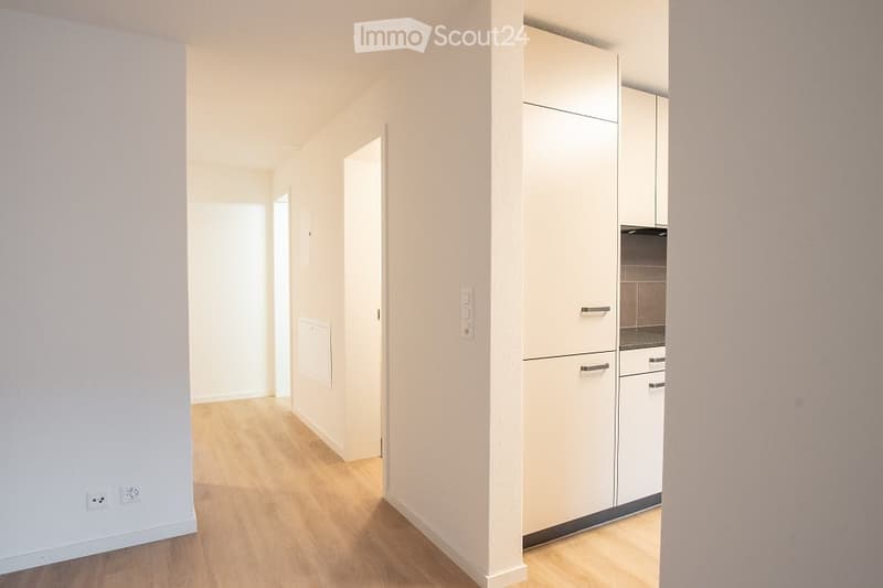Empty hallway with light wooden flooring, white walls, and a small fitted kitchen area visible at the end.