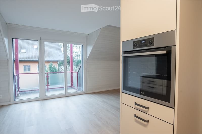 Bright, unfurnished room with wooden flooring, large sliding glass doors leading to a balcony, and a modern kitchen area with a built-in oven.