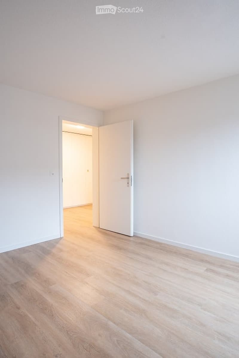 Empty room with light wooden flooring and white walls, featuring an open doorway leading to another room.